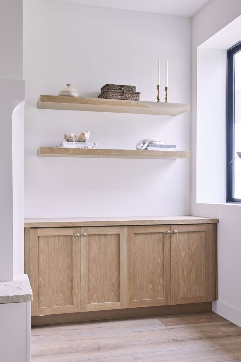 A clean, minimalist built-in featuring two floating white oak shelves styled with books, candles, and decorative objects, above a four-door white oak base cabinet with brushed nickel knobs and a light stone countertop.