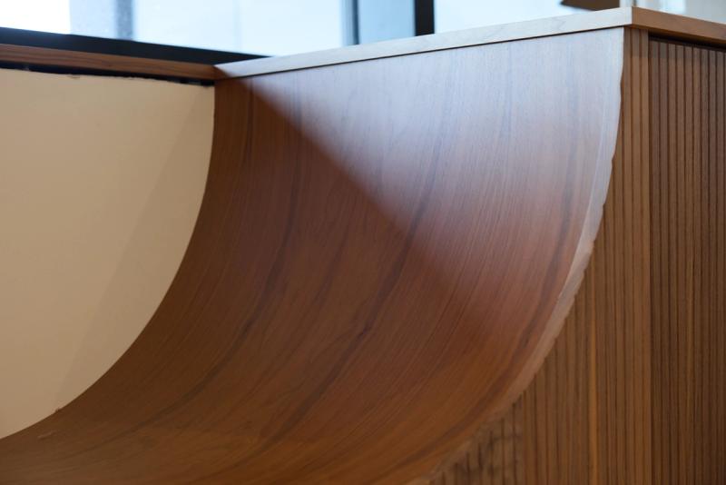 A close-up view of a curved wooden built-in or reception desk featuring smooth bent wood veneer with visible grain and vertical ribbed paneling on the side.