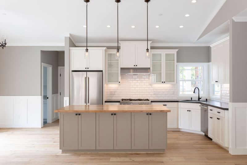 A bright, open kitchen featuring white upper cabinets with glass fronts, a greige shaker-style island with a butcher block countertop, stainless steel refrigerator, white subway tile backsplash, and light hardwood floors. Three pendant lights hang above the island.