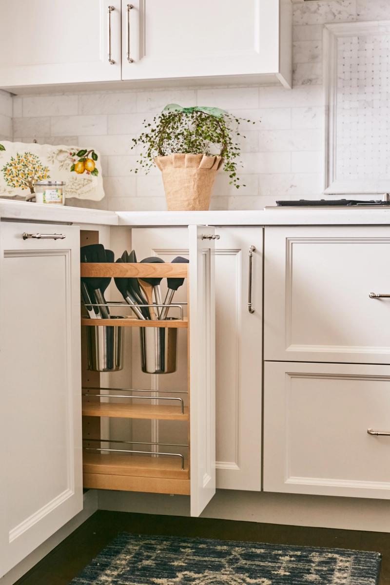 A close-up of a narrow pull-out cabinet organizer between white inset kitchen cabinets, holding stainless steel canisters filled with cooking utensils, with a potted plant and lemon-print dish towel visible on the countertop above.