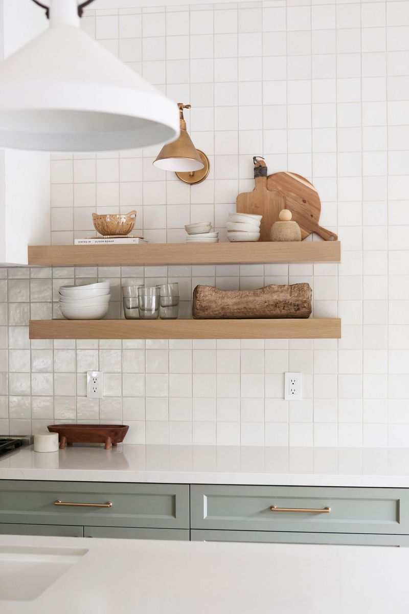 A bright, modern kitchen featuring two floating light oak shelves styled with white bowls, wooden cutting boards, and glassware, set against a white square tile backsplash above sage green lower cabinets with brass hardware.