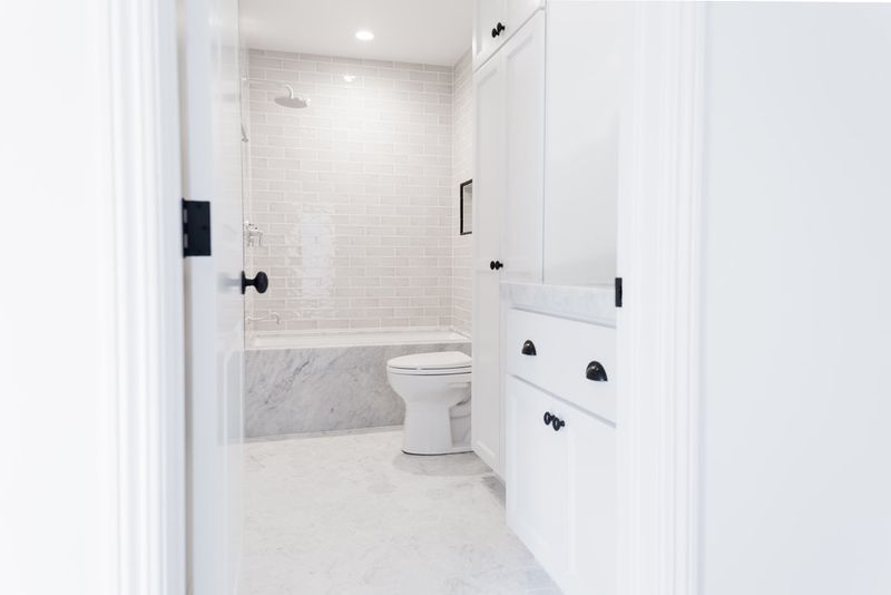 A bright, clean bathroom featuring white shaker-style cabinetry with matte black hardware, marble flooring and tub surround, subway tile walls, and a built-in shower niche.