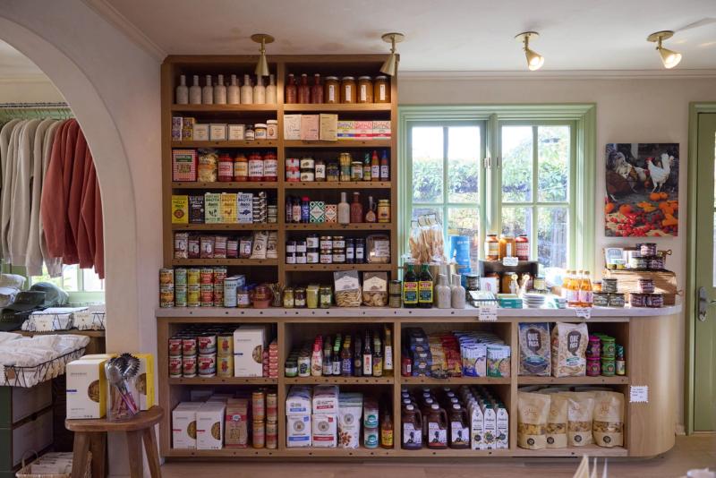 A warmly lit specialty food shop featuring floor-to-ceiling oak shelving units filled with gourmet pantry goods, sauces, and packaged foods, alongside a curved display counter with additional products. Green-trimmed windows allow natural light to fill the space.