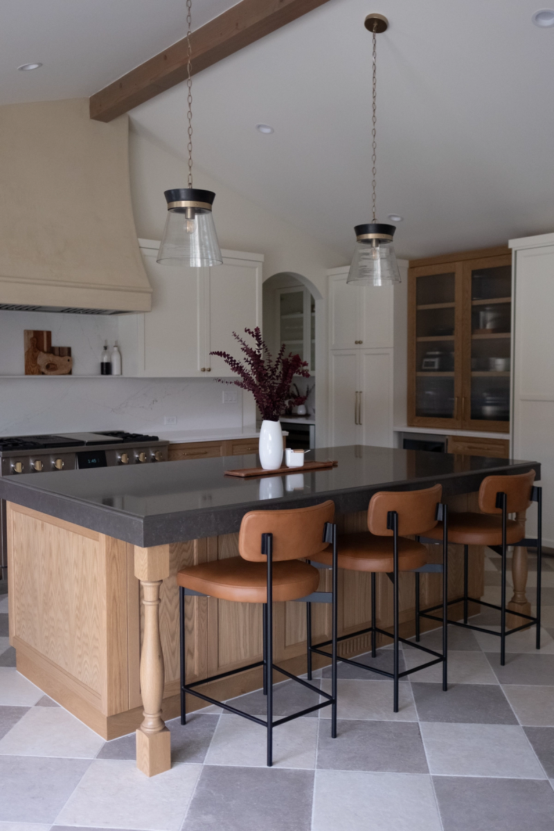 A spacious custom kitchen featuring a large oak island with dark stone countertop, tan leather bar stools, white upper cabinets, oak glass-front display cabinets, and a checkerboard tile floor with two glass pendant lights overhead.
