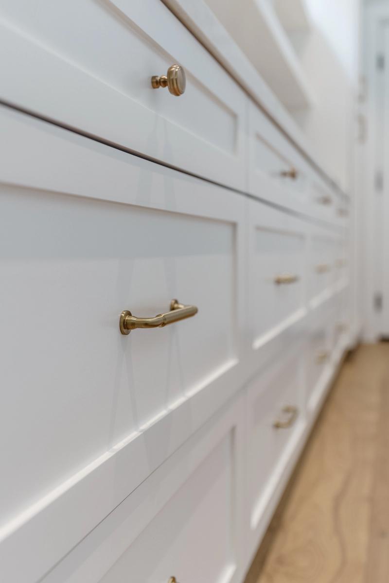 A close-up side-angle view of white custom-built cabinetry with inset panel drawers and brass knob and pull hardware, with warm wood flooring visible in the background.