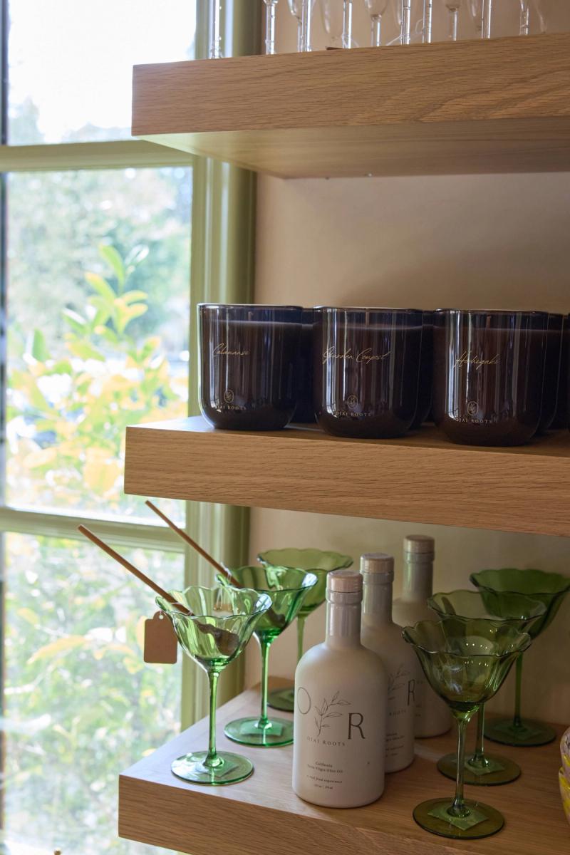 Light oak floating shelves styled with dark amber candles, green martini glasses, and Ojai Roots olive oil bottles in front of a bright window with greenery outside.