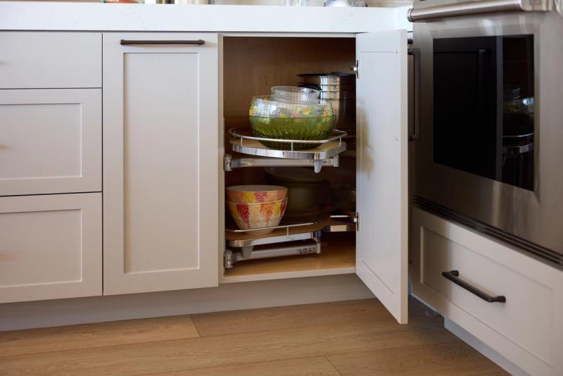 An open corner base cabinet in a white shaker-style kitchen reveals a two-tier lazy Susan organizer holding mixing bowls and glassware, with a stainless steel range visible to the right.