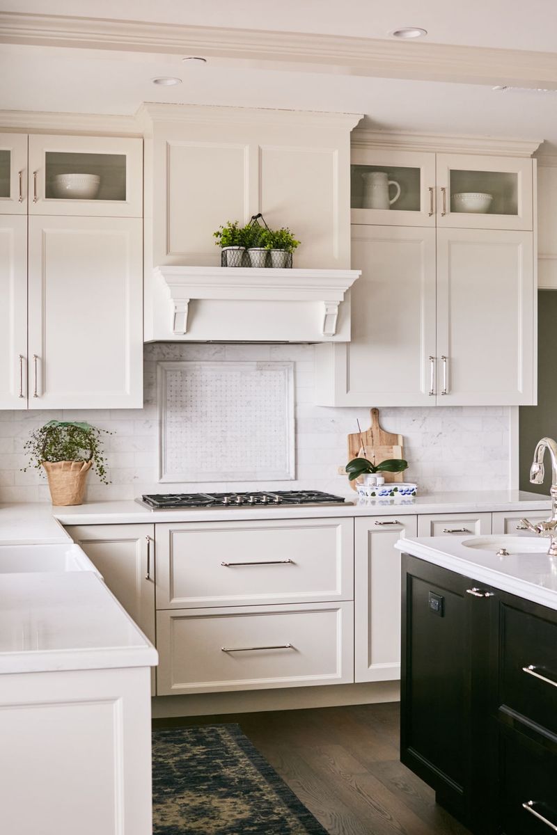 A bright, elegant kitchen featuring custom white inset cabinetry, a decorative wood range hood surround, marble backsplash, gas cooktop, and contrasting dark kitchen island with white quartz countertops.