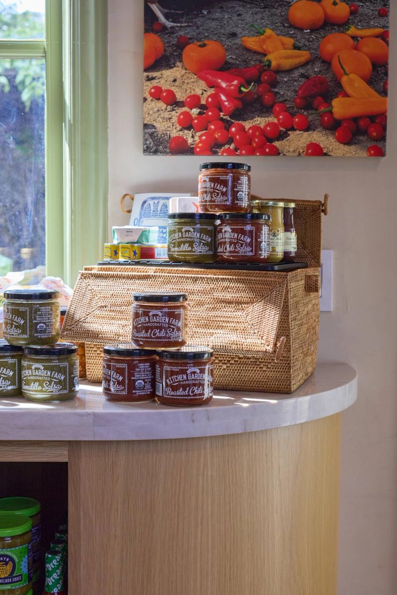 A curved light wood kitchen island with a white marble countertop, styled with handcrafted Kitchen Garden Farm salsa jars in a wicker basket display, with a vegetable photograph on the wall behind.
