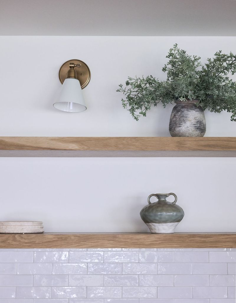 Two floating white oak shelves mounted on a white wall above white subway tile, styled with a textured ceramic vase holding greenery, a two-toned ceramic jug, and a woven dish, alongside a brass wall sconce.