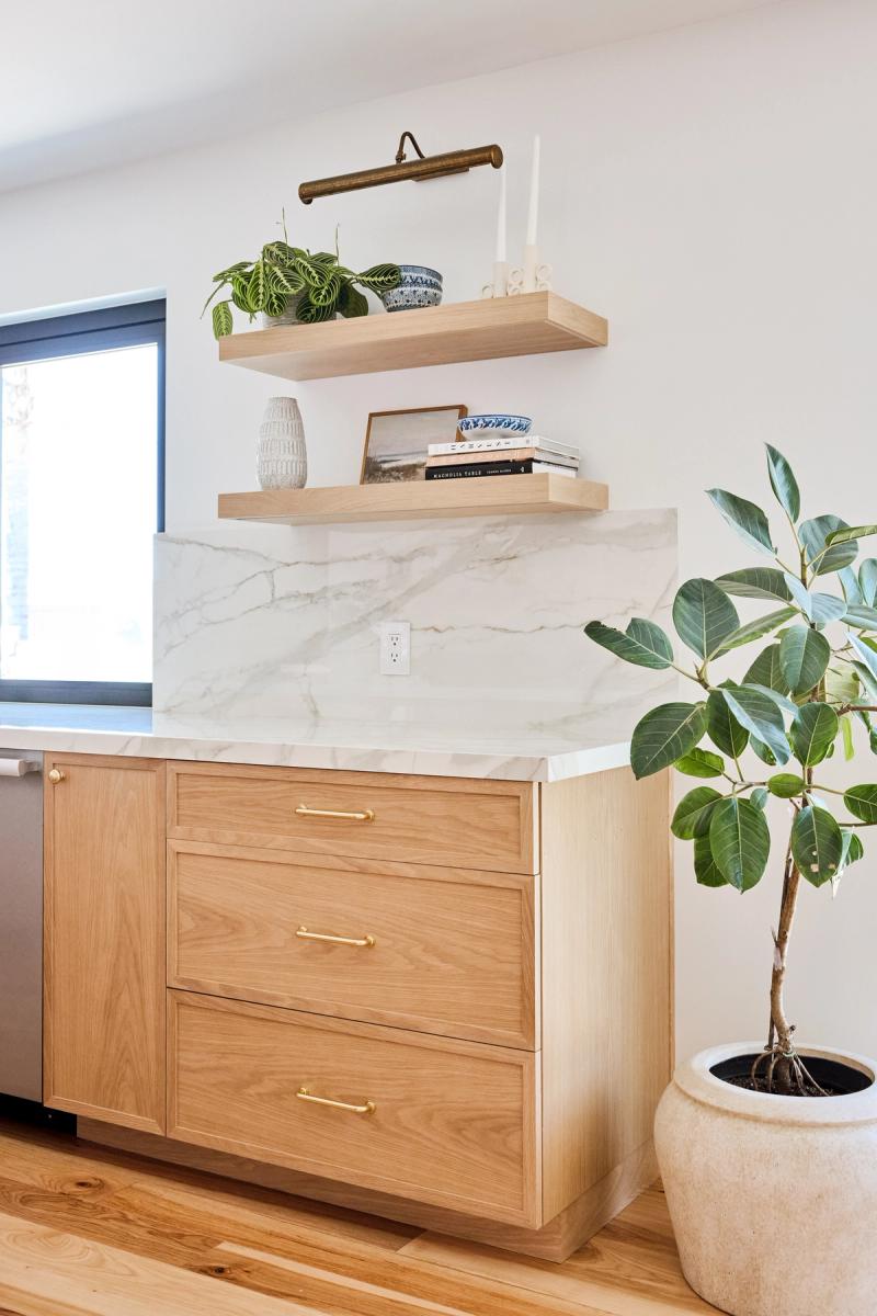 A modern kitchen featuring white oak cabinetry with brass hardware, a white marble countertop and backsplash, two floating wood shelves decorated with plants and blue pottery, and a large potted fiddle leaf fig tree in the foreground.