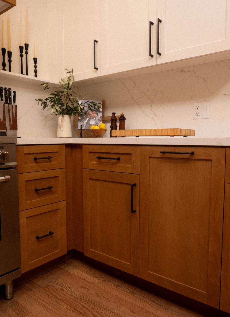 A kitchen featuring warm oak lower cabinets paired with cream upper cabinets, both with matte black hardware, set against a white marble-look quartz backsplash and countertop. The counter is styled with candle holders, a plant, lemons, and a wooden cutting board.