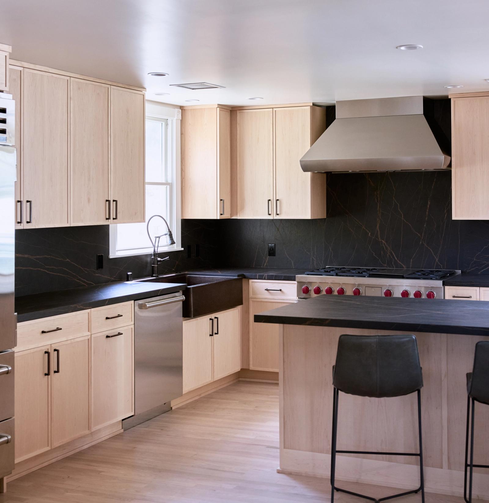 A modern custom kitchen featuring light maple flat-panel cabinets, dramatic black soapstone countertops and backsplash, a copper farmhouse sink, professional-grade range with red knobs, stainless steel range hood, and a center island with dark leather barstools.