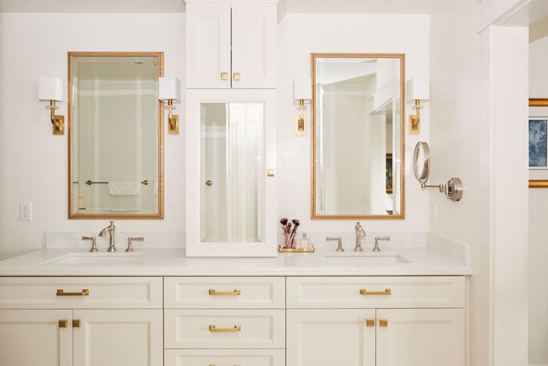 A bright white bathroom vanity with two undermount sinks, brushed nickel faucets, gold cabinet hardware, and gold-framed mirrors flanked by brass wall sconces. White shaker-style cabinetry with a marble-look countertop fills the full width of the wall.