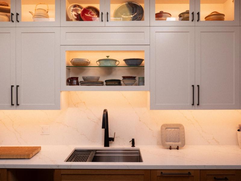 A modern kitchen featuring light gray shaker-style cabinets with black hardware, an open illuminated display shelf showcasing handmade pottery, a marble-look quartz backsplash, and a black undermount sink with pull-down faucet.