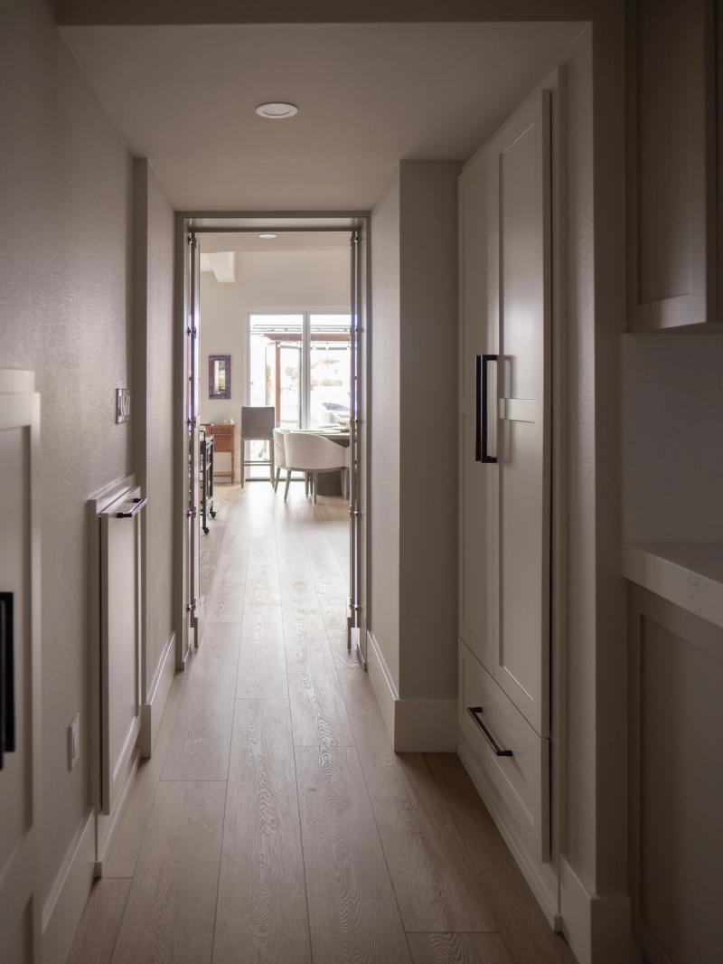 A well-lit residential hallway featuring light wood flooring and custom gray-toned cabinetry with matte black bar pulls, leading through glass-paneled doors into a bright dining area.