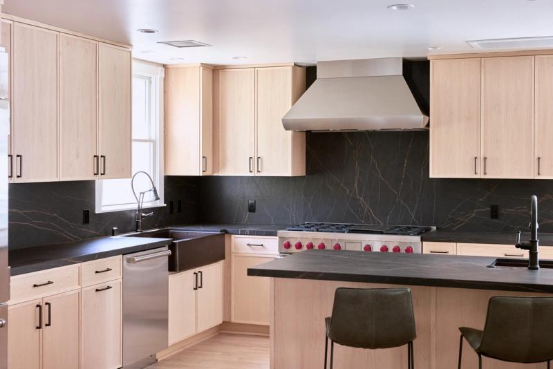 A modern kitchen featuring light maple flat-panel cabinetry, dark soapstone countertops and backsplash, a professional stainless range with red knobs, and an island with bar seating.