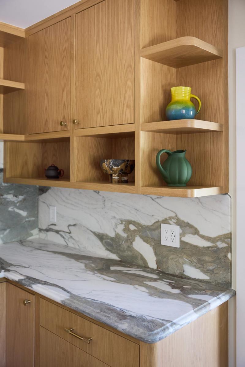 A detail view of a custom white oak kitchen featuring open shelving with decorative ceramic pitchers, closed upper cabinets with brass hardware, and a dramatic book-matched marble countertop and backsplash with green and grey veining.
