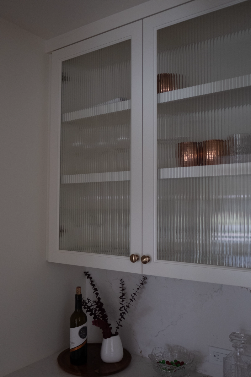 White upper kitchen cabinets with vertical reeded glass panel doors and brushed brass knobs, displaying copper glassware inside. Below the cabinets sits a marble countertop with a wine bottle, dried eucalyptus in a small vase, and decorative bowls.