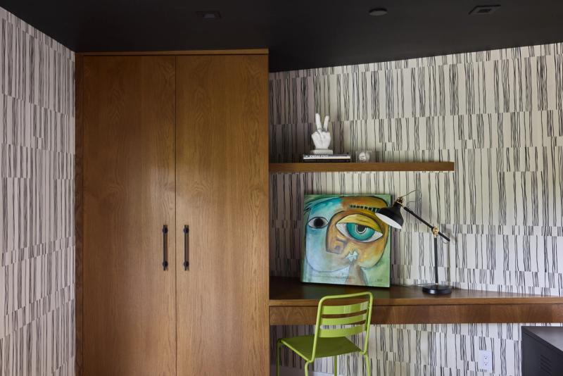 A stylish home office nook featuring warm oak built-in floating desk and wall cabinet with black hardware, paired with bold graphic wallpaper, a lime green chair, and eclectic artwork.