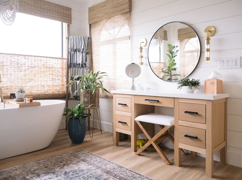 A bright, airy bathroom featuring a natural oak vanity with black hardware and white marble top, paired with a round black-framed mirror and gold sconce lighting. A freestanding soaking tub and woven bamboo shades are visible in the background.