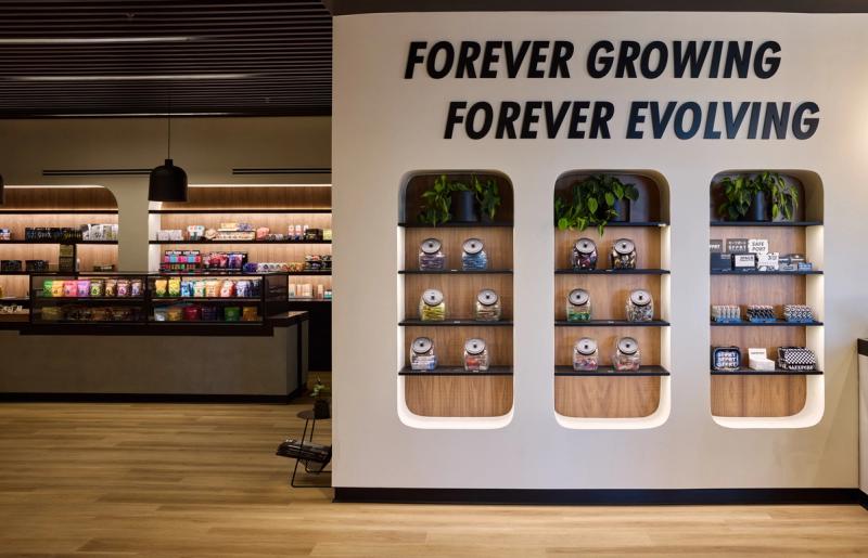 A modern retail interior featuring three arched built-in wood and dark shelving units displaying glass jars and packaged products, with a 'Forever Growing Forever Evolving' slogan on the wall above. A product display counter with colorful packaging is visible in the background.