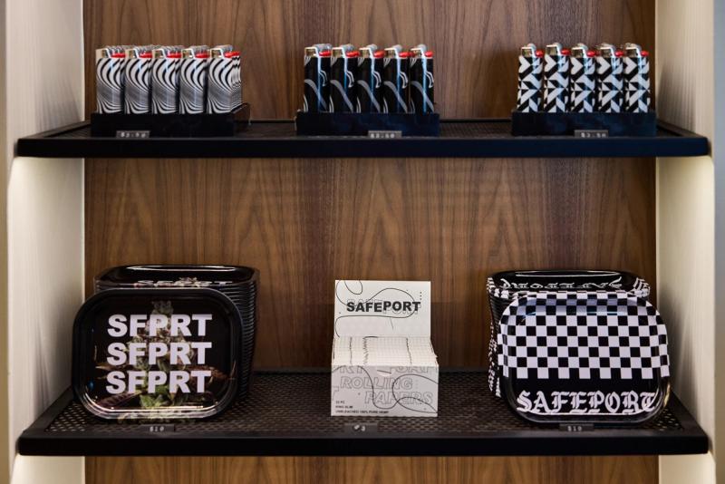 A built-in wood shelving unit with black metal mesh shelves displaying branded lighters, rolling trays, and rolling papers for a retail store called Safeport.