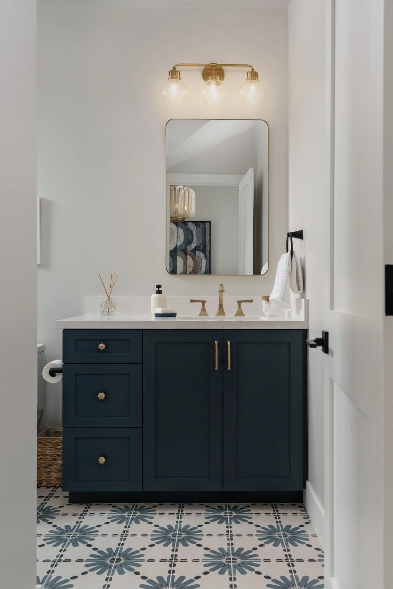 A custom dark navy blue shaker-style bathroom vanity with brass fixtures, gold-framed mirror, and decorative patterned floor tile, photographed from a hallway doorway.