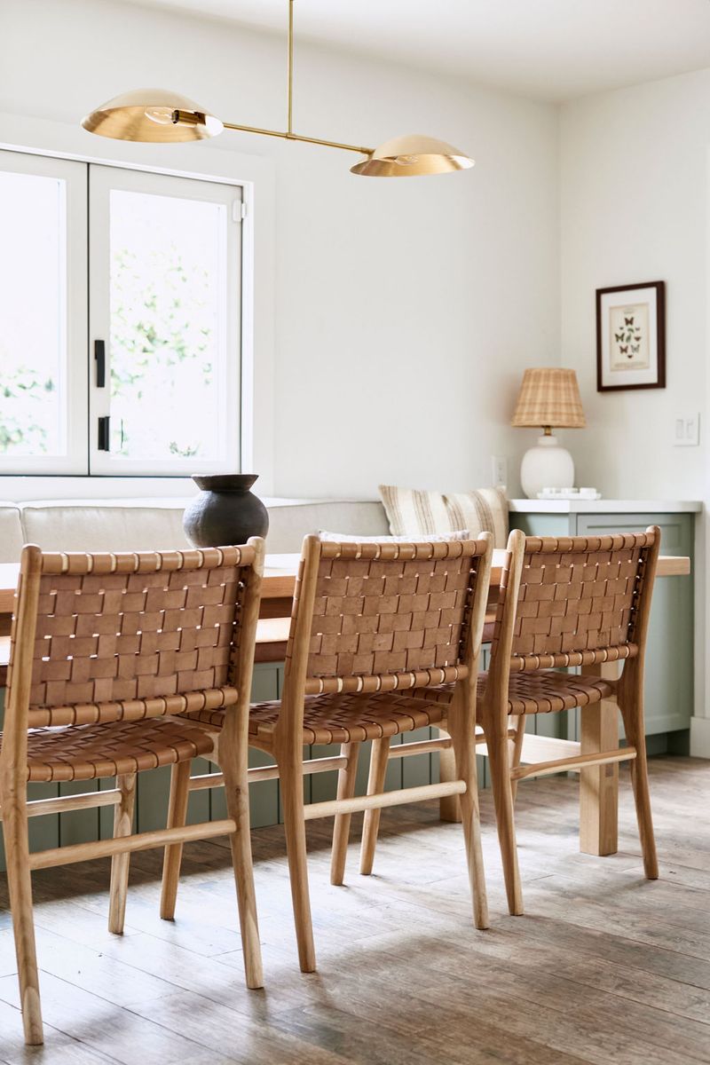 A bright dining room featuring woven leather chairs around a wood table, with a sage green built-in sideboard cabinet along the back wall, topped with a white ceramic lamp and framed botanical art.