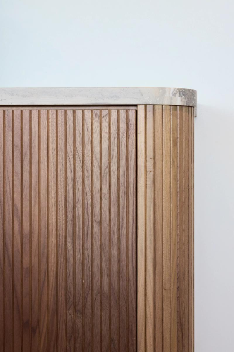 A close-up of a custom wood cabinet featuring vertical fluted panels in warm walnut tones, a rounded corner profile, and a stone or travertine top surface.