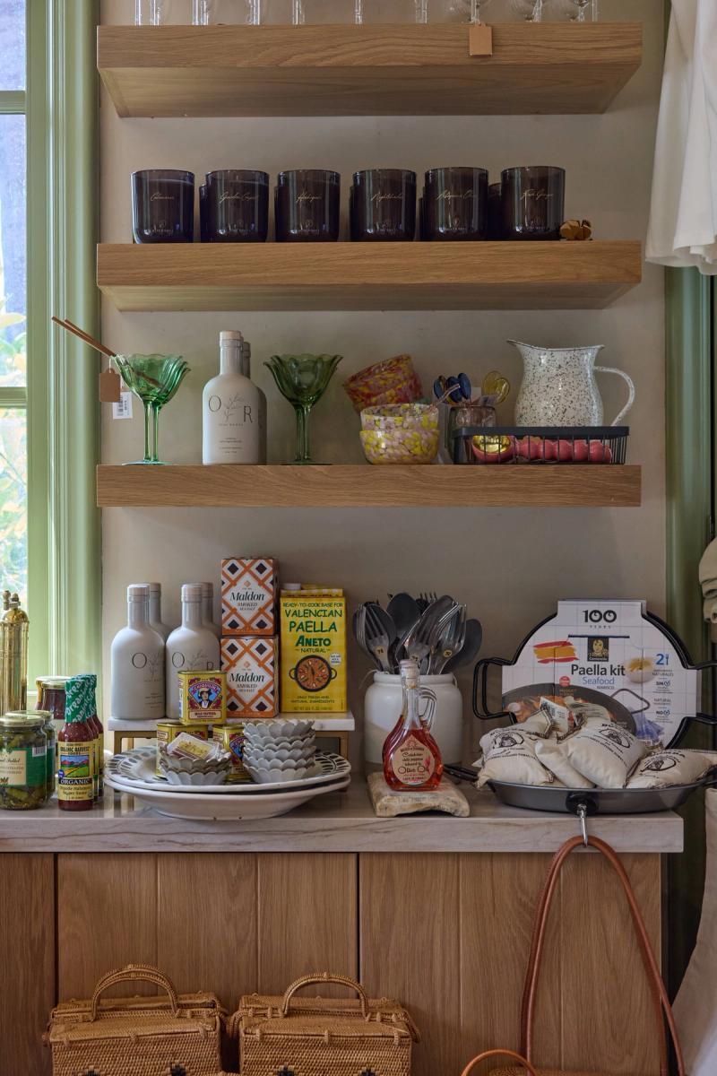 A styled kitchen vignette featuring light oak floating shelves mounted on sage green walls, displaying candles, glassware, olive oil bottles, and gourmet food products including a paella kit and Maldon salt.
