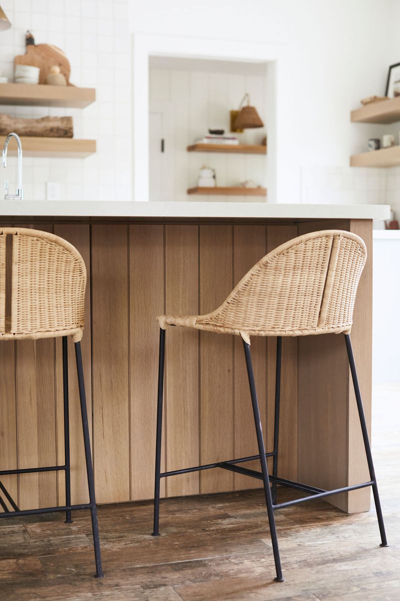 Two wicker and black metal bar stools sit at a white oak kitchen island with a white quartz countertop, surrounded by floating wood shelves and white tile backsplash.