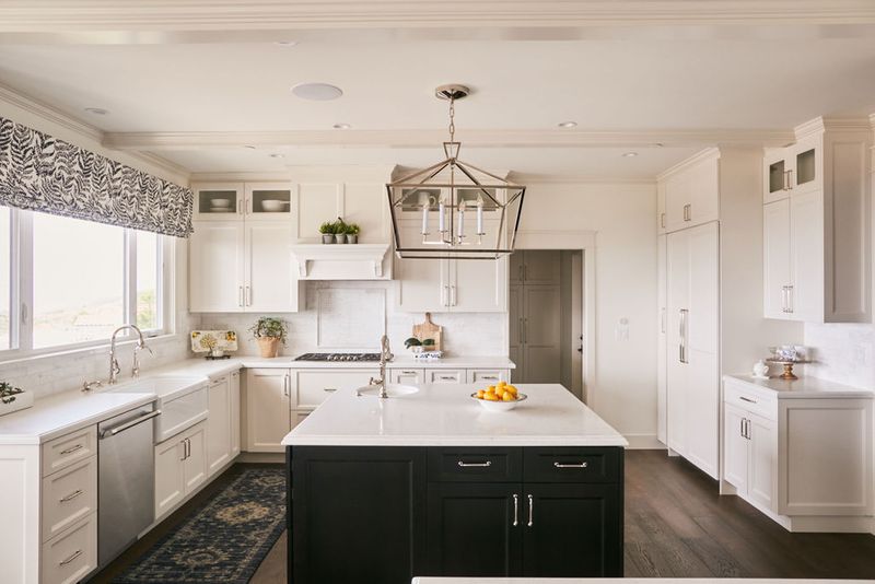 A bright, transitional-style kitchen featuring white shaker cabinets, a dark navy island with white quartz countertop, geometric pendant light, and dark hardwood floors.
