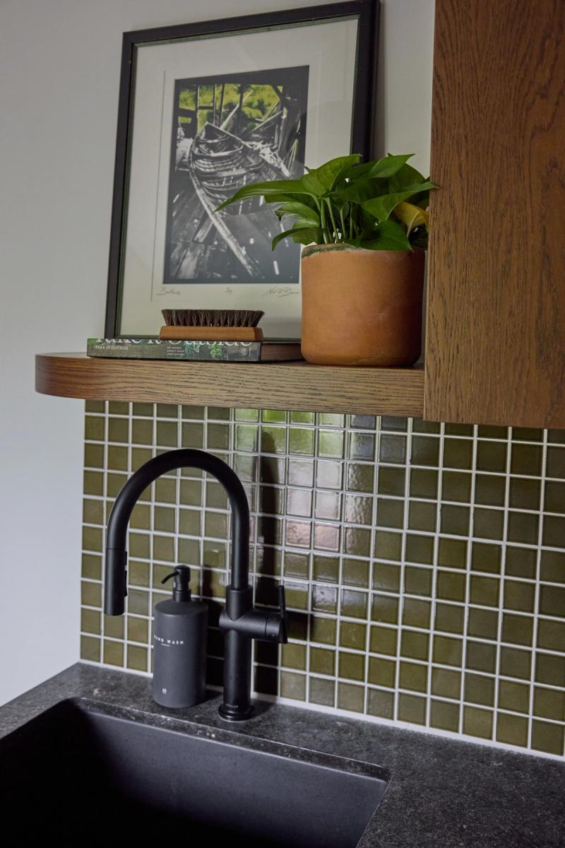 A close-up of a kitchen or utility sink area featuring olive green mosaic tile backsplash, a matte black gooseneck faucet, a floating oak shelf with a potted plant, small brush, and book, and a framed black and white print.