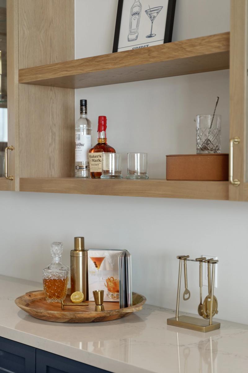 A styled home bar area featuring light oak open shelving with brass hardware, holding whisky and vodka bottles and glassware, above a white quartz countertop with a wooden tray, gold cocktail shaker, and bar tools.