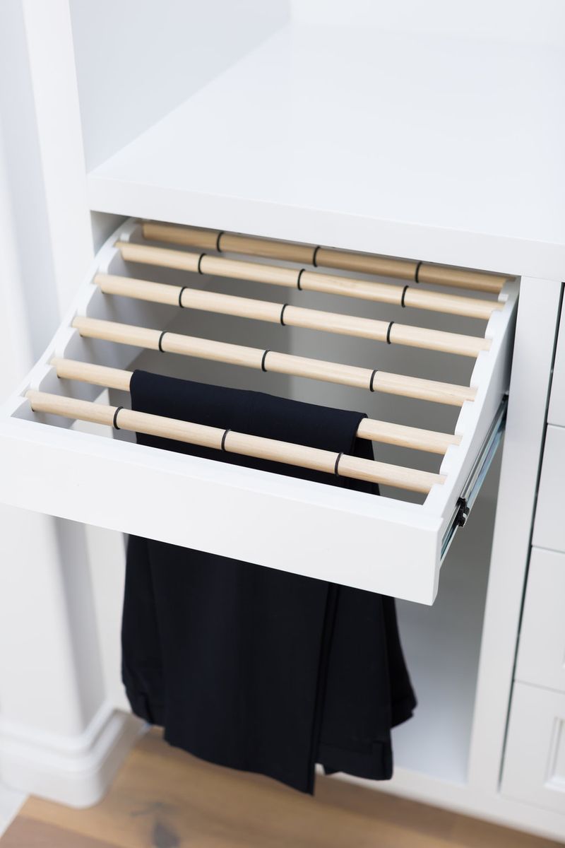 A white pull-out drawer fitted with multiple wooden dowel rods for hanging trousers, with a pair of black pants draped over one of the rods, set inside a white custom closet cabinet.