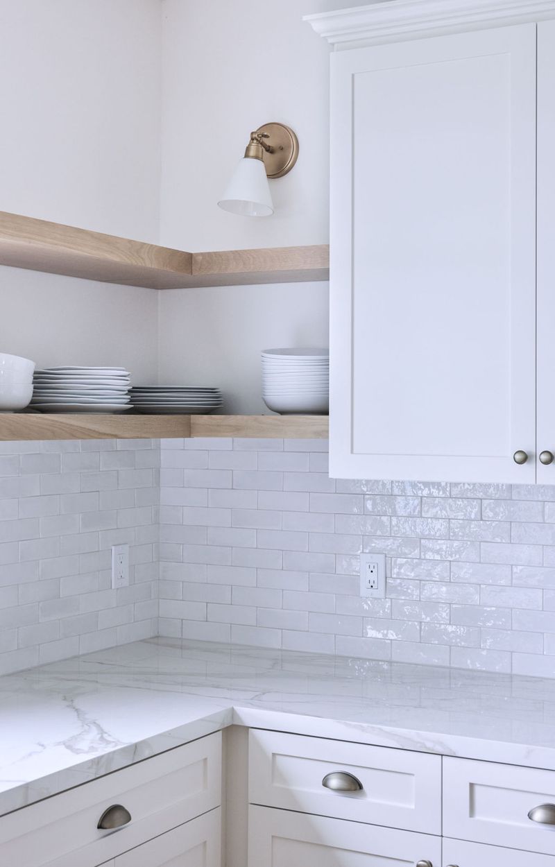 A bright, clean kitchen corner featuring white shaker cabinets, light wood open shelving stacked with white dishes, a white subway tile backsplash, marble countertops, and a brass wall sconce.