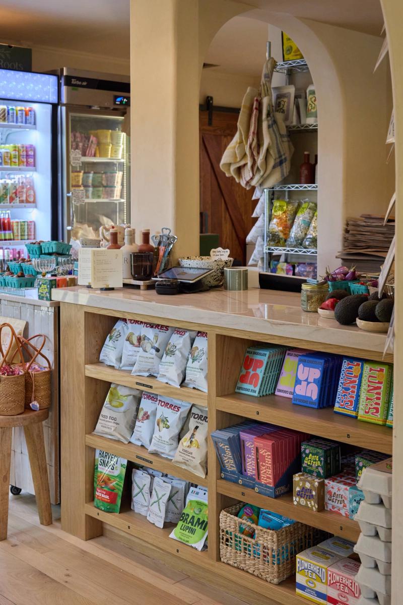 A light wood built-in counter and open shelving unit in a boutique retail shop, stocked with specialty snacks, chocolates, and teas, with an arched architectural detail and a beverage refrigerator visible in the background.