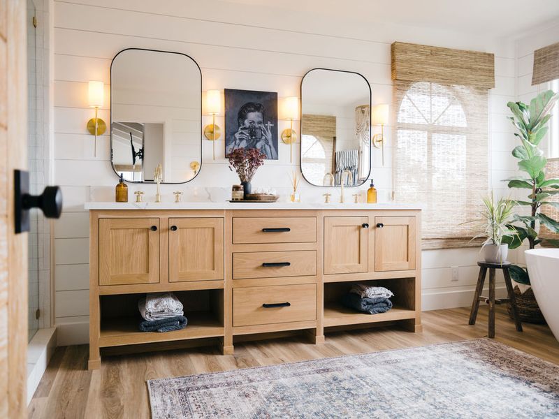 A spacious master bathroom featuring a wide natural oak double vanity with black hardware, white marble countertops, dual rounded mirrors with gold sconce lighting, and open shelving with folded towels. A freestanding tub and fiddle leaf fig tree are visible to the right.