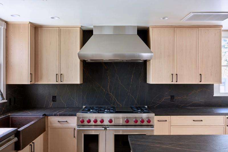 A modern kitchen featuring flat-panel natural maple cabinets, dramatic black and gold veined soapstone countertops and full-height backsplash, a professional stainless steel range with red knobs, and a stainless range hood.