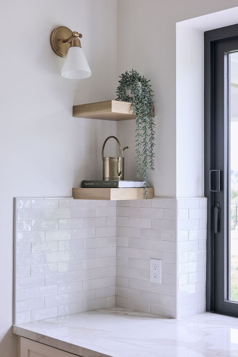 A bright kitchen corner featuring glossy white subway tile backsplash, light wood floating shelves styled with books and a trailing plant, a brass wall sconce, and a marble countertop beside a black-framed glass door.