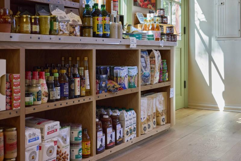 A well-lit retail interior featuring custom natural oak shelving units stocked with pantry goods, oils, and dry goods. The cabinetry displays clean joinery and a light wood finish complemented by a green-painted door in the background.