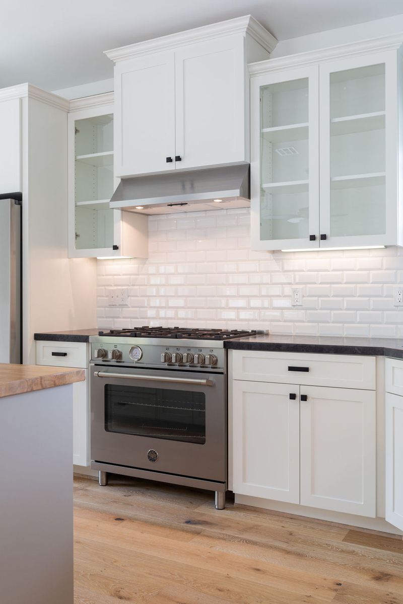 A bright white shaker-style kitchen featuring a professional stainless steel gas range, subway tile backsplash, dark stone countertops, glass-front upper cabinets, and light hardwood flooring.