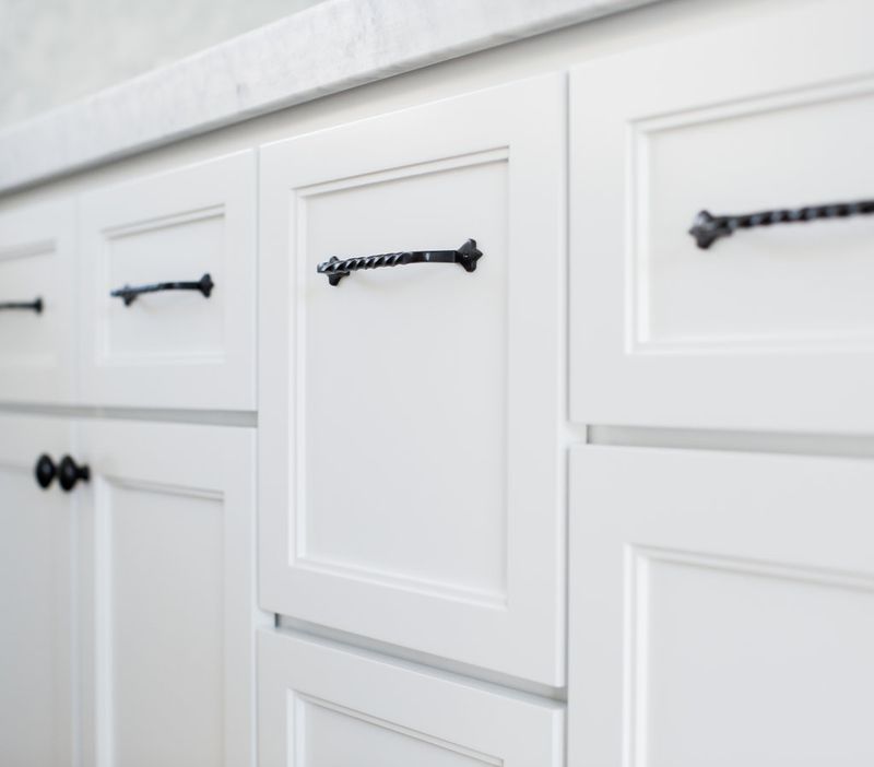 Close-up of white painted inset bathroom vanity cabinets with decorative twisted black iron bar pulls on drawers and round knobs on cabinet doors, topped with a white marble countertop.