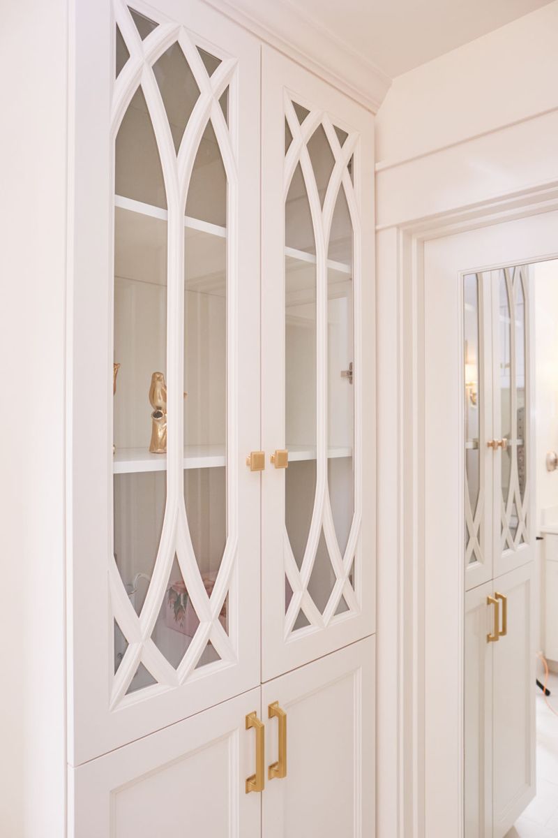 Elegant white built-in cabinetry with gothic arch lattice glass doors and brushed brass hardware, featuring open display shelves with gold decorative accents.
