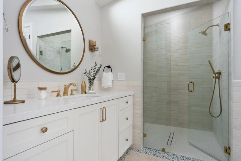 A bright, modern bathroom featuring a white shaker-style vanity with brass hardware, a round gold-framed mirror, and a glass-enclosed walk-in shower with sage green tile and brushed brass fixtures.