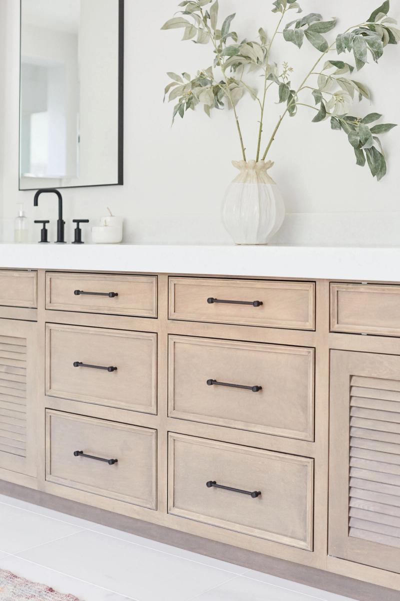 A wide bathroom vanity with light natural wood drawers, black bar pulls, a white quartz countertop, black faucet, and a white ceramic vase with eucalyptus branches.
