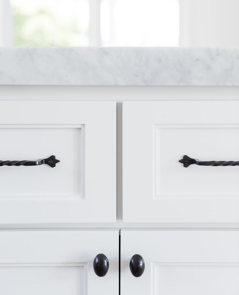 Close-up of white shaker-style cabinet drawers and doors with twisted black iron pull handles and oval knobs, topped with a gray marble countertop.