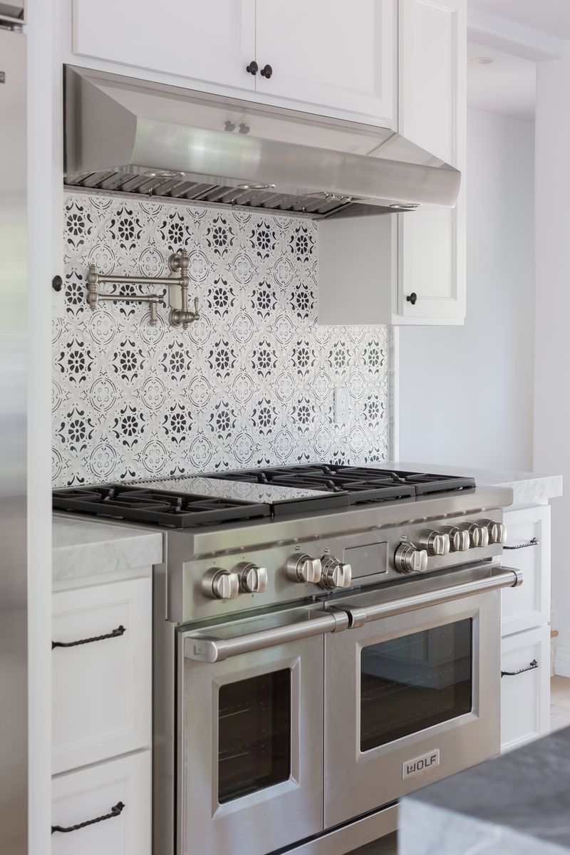 A stainless steel Wolf dual-oven range with six burners set into white custom cabinetry, featuring a decorative black and white patterned tile backsplash and stainless steel range hood.