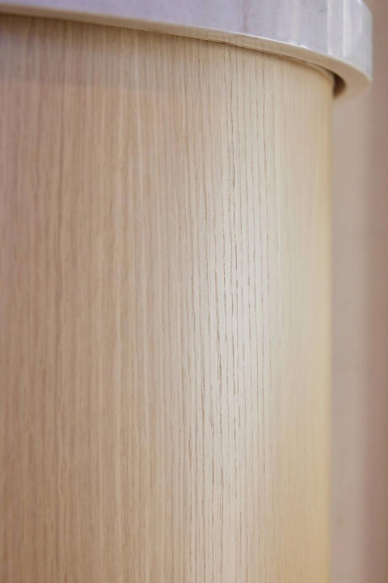 A close-up detail shot of a curved white oak wood panel or cabinet face, showing fine vertical grain texture and a smooth rounded edge with a light-colored trim at the top.